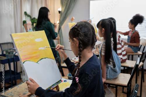A group of multiracial kids learning with a female Asian teaches acrylic color picture painting on canvas in art classroom, creatively learning with skill at the elementary school studio education. photo