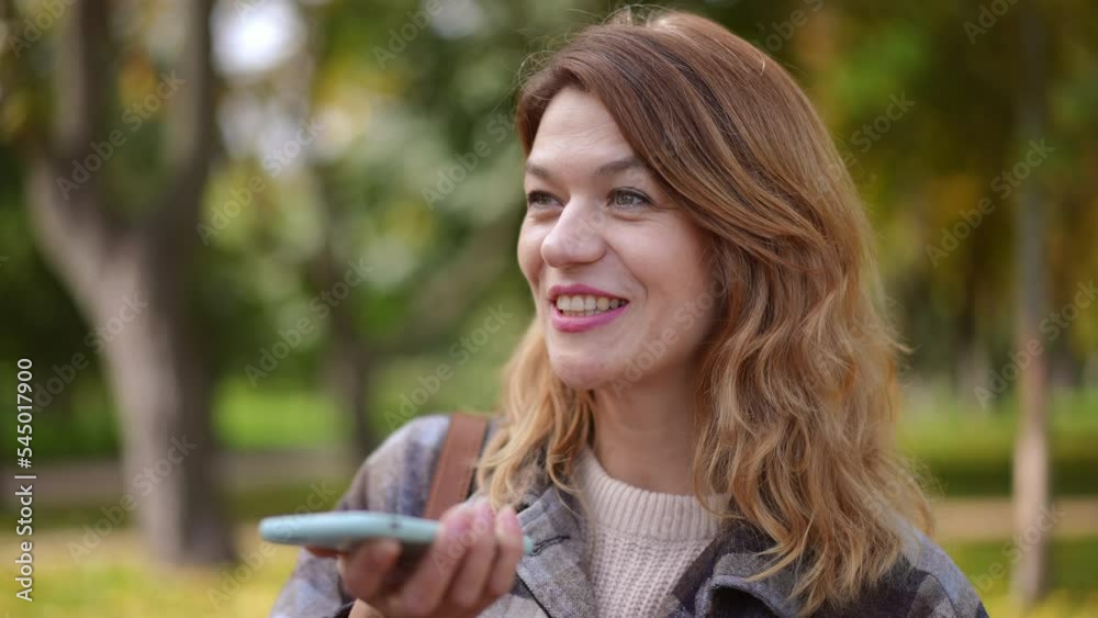 Positive brunette woman with hazel eyes talking on smartphone loudspeaker smiling standing in autumn park. Portrait of confident charming Caucasian lady chatting in slow motion on sunny day outdoors Positive brunette woman with hazel eyes talking on smartphone loudspeaker smiling standing in autumn park. Portrait of confident charming Caucasian lady chatting in slow motion on sunny day outdoors