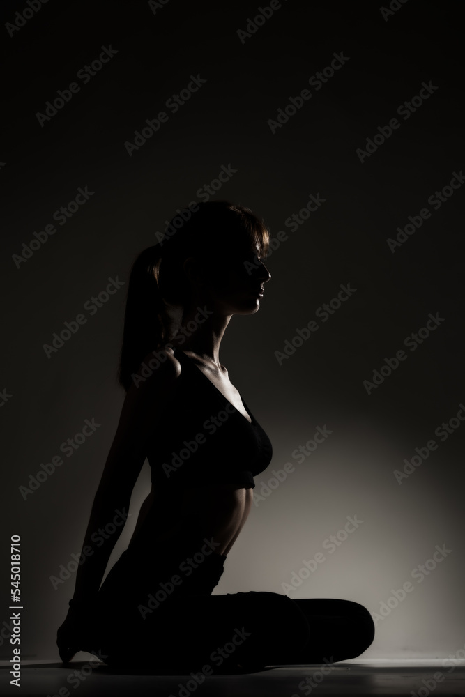 Fit girl resting and stretching after practicing yoga poses. Side lit half silhouette against dark background. .