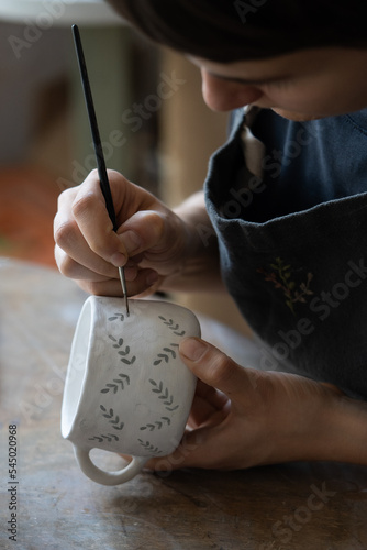 Concentrated woman potter holding brush and painting minimalistic pattern on clay cup. Attentive girl in apron sits at table in workshop carefully applies ornament to vessel enjoying creative process