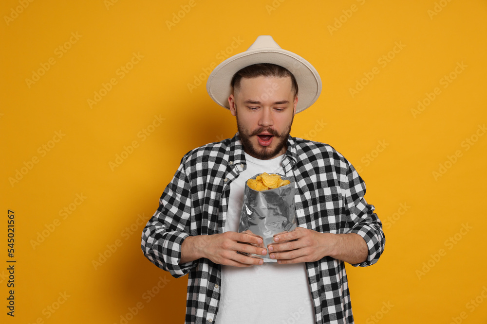 Emotional young man with bag of tasty potato chips on yellow background Emotional young man with bag of tasty potato chips on yellow background