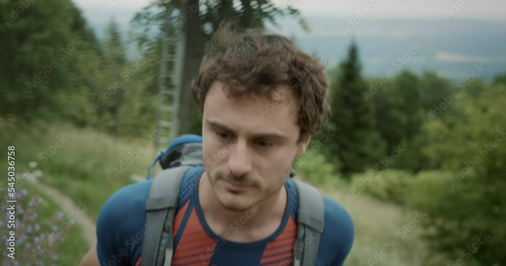 Close up of a young hiker walking up a narrow road towards the peak of a mountain. Walking past the trees and flowers, with hands holding the straps of the backpack. Close up of a young hiker walking up a narrow road towards the peak of a mountain. Walking past the trees and flowers, with hands holding the straps of the backpack.