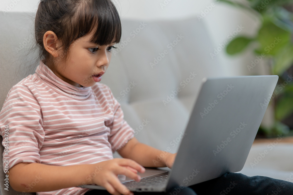 Asian Little school kid girl use laptop computer sitting on sofa alone at home. Child learning reading online social media content, play education lessons game chatting with friends. Asian Little school kid girl use laptop computer sitting on sofa alone at home. Child learning reading online social media content, play education lessons game chatting with friends.
