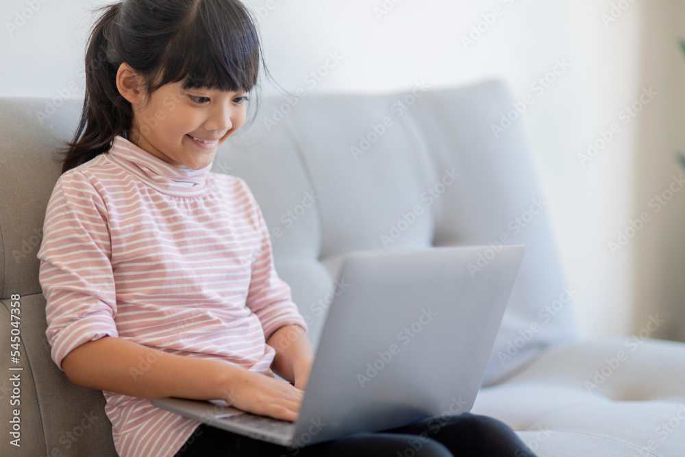 Asian Little school kid girl use laptop computer sitting on sofa alone at home. Child learning reading online social media content, play education lessons game chatting with friends. Asian Little school kid girl use laptop computer sitting on sofa alone at home. Child learning reading online social media content, play education lessons game chatting with friends.