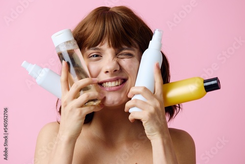 a close photo on a pink background of a funny beautiful woman standing with different jars of care cosmetics, bringing them close to her face and smiling broadly, squinting her eyes in a funny grimace