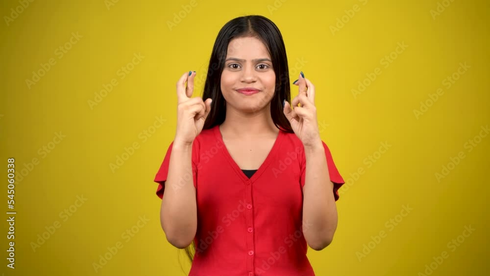Portrait of a smiling young girl with long hair standing over yellow background, holding fingers crossed for good luck Portrait of a smiling young girl with long hair standing over yellow background, holding fingers crossed for good luck