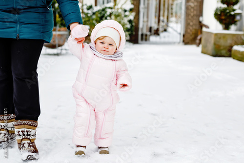 Adorable little baby girl making first steps outdoors in winter through snow. Cute toddler learning walking. Mother holding child on hand. Daughter and mum walk together. Happy family