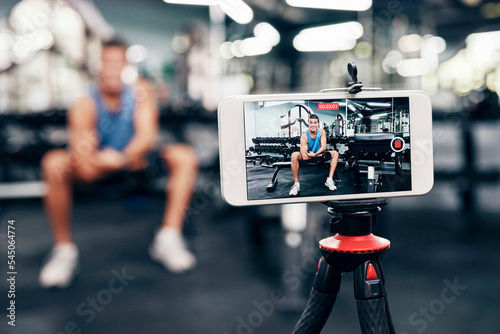 Fototapeta Naklejka Na Ścianę i Meble -  Gym, social media and fitness influencer with phone live streaming workout for interactive multimedia broadcast. Vlog, man filming arm exercise and training coach video recording online blog tutorial