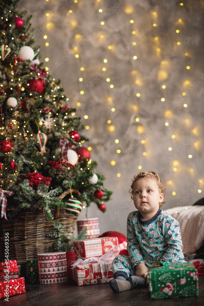 A little one and a half year old boy in pajamas sits under the Christmas tree and plays with a box with gifts. bokeh in the background from the New Year's garland A little one and a half year old boy in pajamas sits under the Christmas tree and plays with a box with gifts. bokeh in the background from the New Year's garland