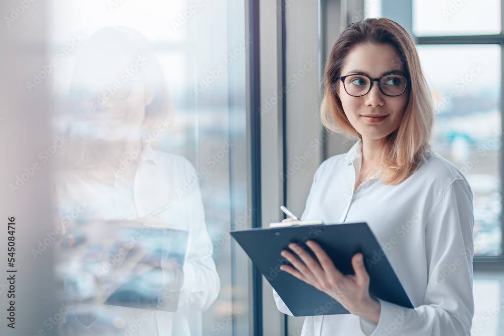 Young woman standing at the panoramic window of a modern office with a folder of documents and a pen.