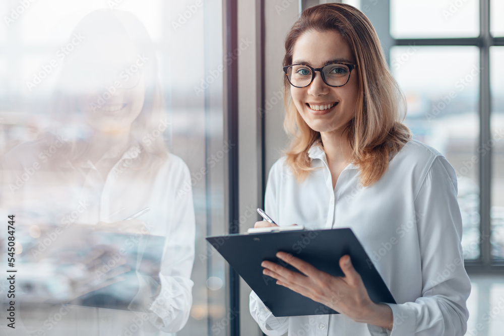 Young smiling woman standing at the panoramic window of a modern office and holding a folder with documents.