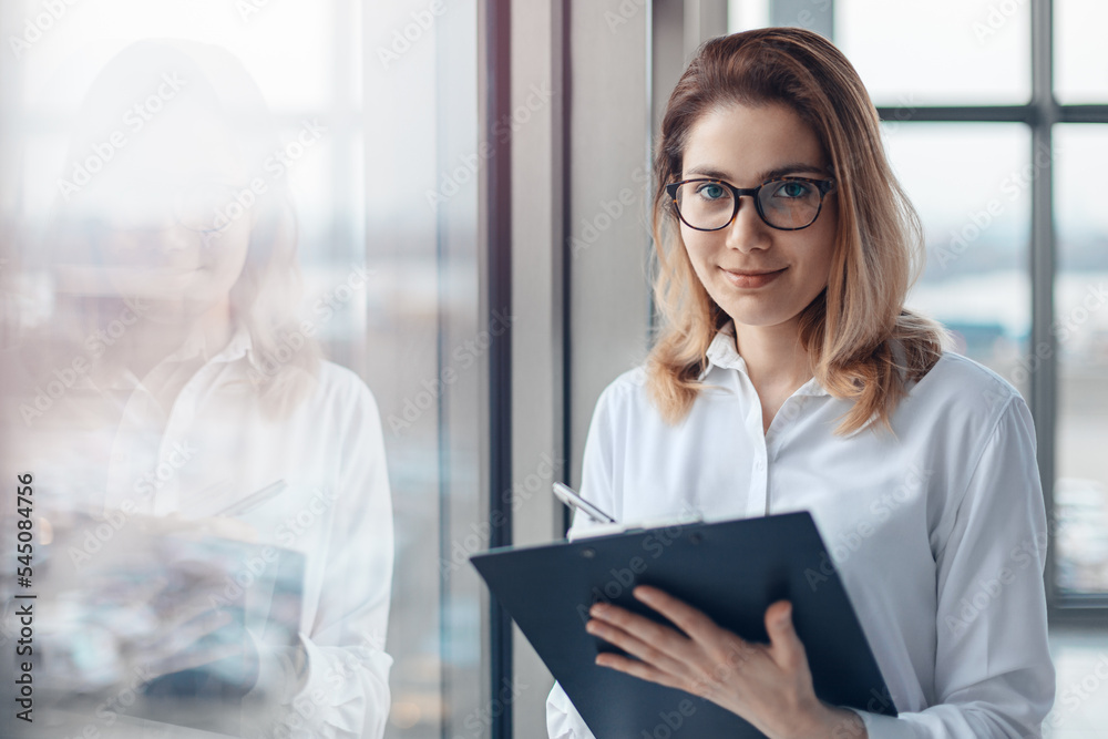 Young business woman standing at the window of a modern office and holding a folder with documents.