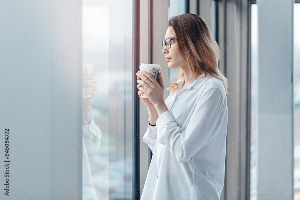 Young woman standing at the window of a modern office and holding a cup of coffee.
