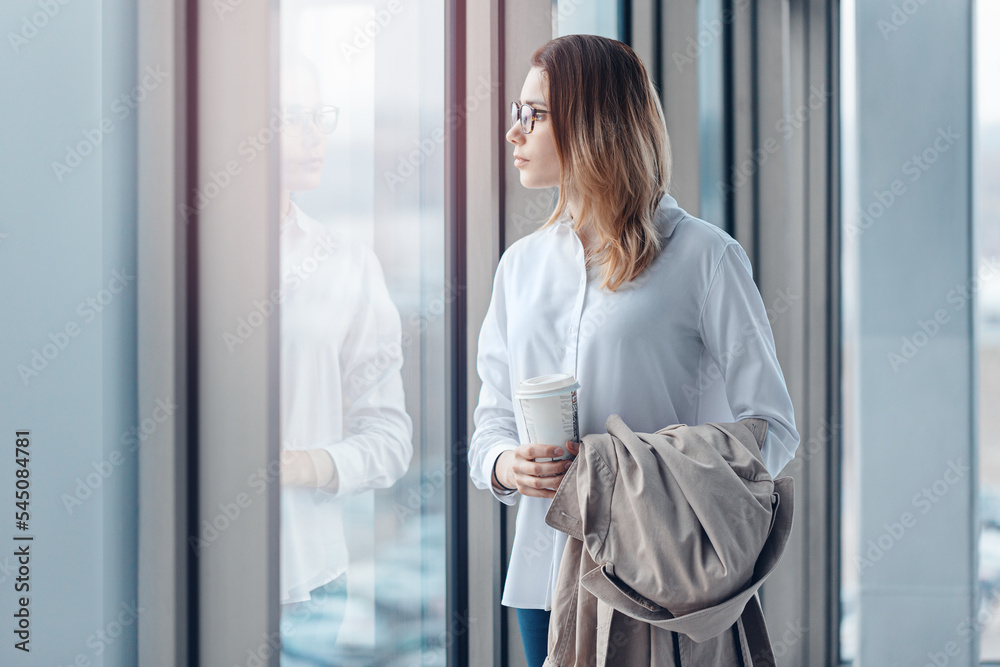 Young woman standing at the window of a modern office with a cup of coffee.