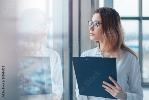 Obraz na płótnie Young woman looking in the panoramic window of a modern office and holding a folder with documents. w salonie