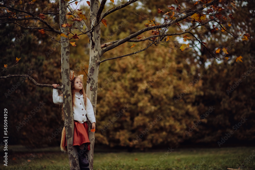 beautiful little girl in a fox costume climbed a tree with yellow orange falling leaves in the park, the child plays, children's leisure, vacations and outdoor activities. children walking in autumn