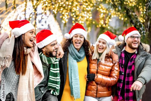 Young friends group walking under Christmas tree decorations - Winter life style concept with trendy happy people wearing red santa hats while having fun holidays together outside - Warm bright filter