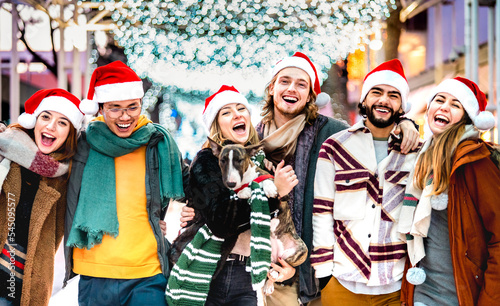 Genuine friends group having fun together with cute dog on Christmas winter advent - Xmas life style concept with trendy young people wearing red santa claus hats out side - Bright vivid neon filter