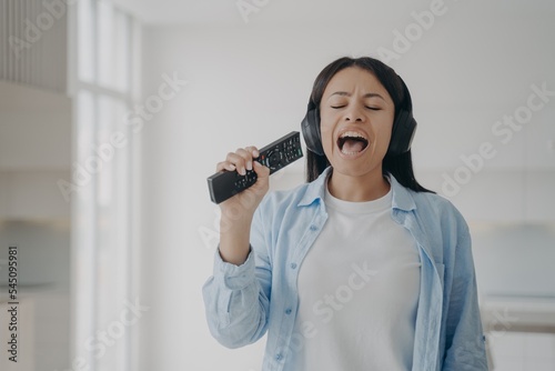 Female singing karaoke, using TV remote control like a mic, listening to music in headphones at home