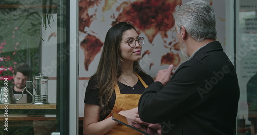 Male business manager managing coffee shop speaking with female employee in front of cafe place holding tablet. Mature entrepreneur talking explaining protocol