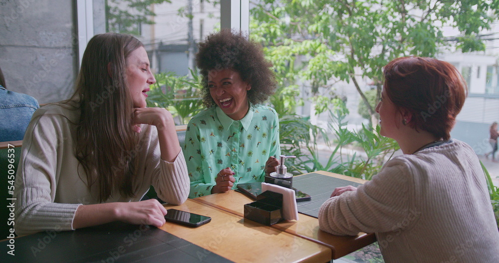 Three happy diverse women sitting at coffee shop table by window overlooking city sidewalk. Group of female friends chatting and laughing in conversation. Authentic real life laugh and smile