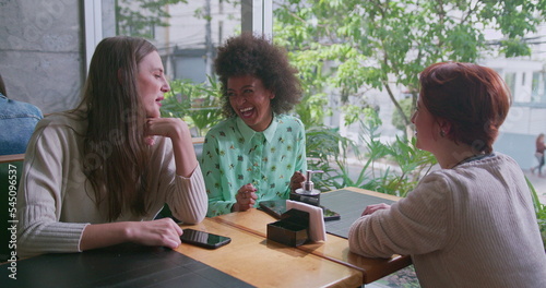 Three happy diverse women sitting at coffee shop table by window overlooking city sidewalk. Group of female friends chatting and laughing in conversation. Authentic real life laugh and smile
