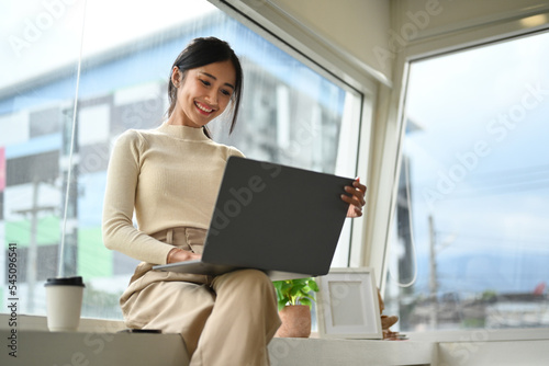Attractive young creative woman using laptop while sitting near office window with cityscape on background