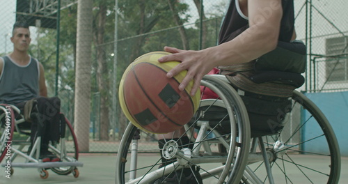 A paraplegic male athlete receiving ball from colleague outdoors. Two disabled athletes playing basketball in sport court outside. Sport and disability concept