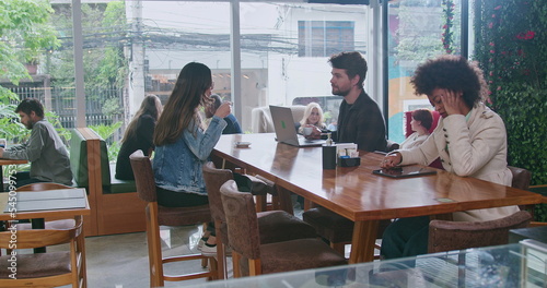 Group of people inside coffee shop. Female friend handing espresso to male colleague sitting at cafe restaurant table. Men and women customers together at cafe place