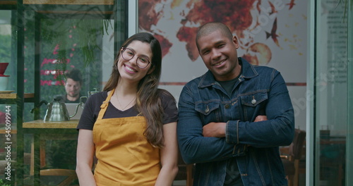Two diverse Brazilian hispanic employees of coffee shop portrait faces smiling. A black manager with a South American female staff standing in front of restaurant entrance