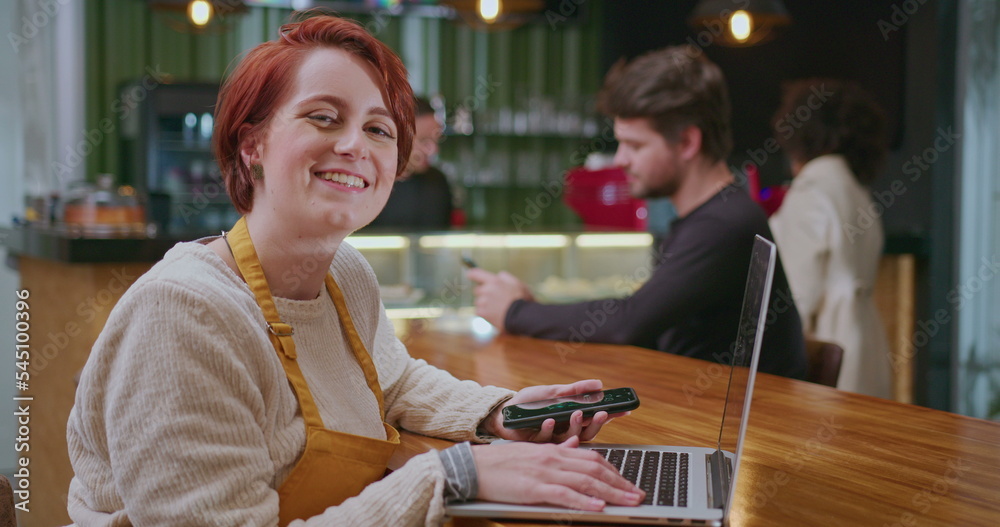 One happy female barista seated at coffee shop table holding phone in front of laptop computer turns head to camera smiling