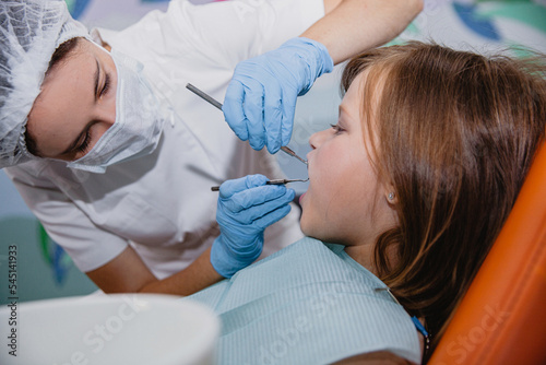 A woman dentist in a white coat and a medical cap treats the teeth of European appearance girl in a dental office