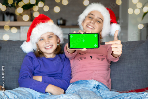 Selective focus on the green smartphone screen in the hands of two cute girls sisters in a Santa hat. A place for advertising, text, greetings. Laughter, joy, good mood.