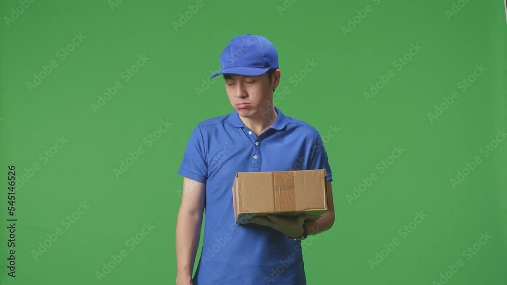 Tired Asian Male Courier In Blue Uniform Shaking His Head, Touching His Face, And Stretching While Delivering A Carton On Green Screen Background In The Studio
