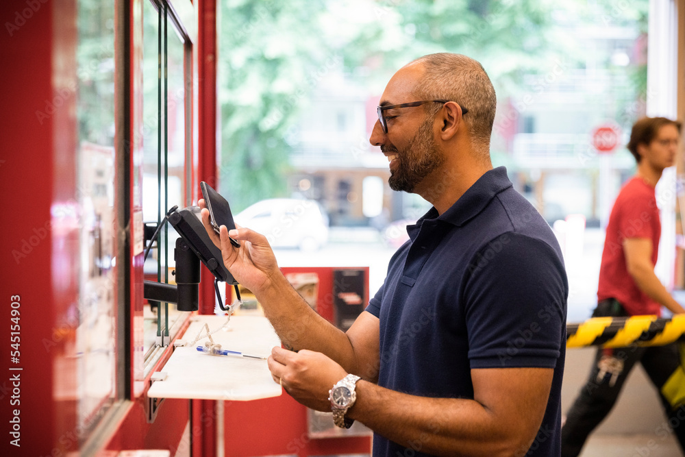 Side view of smiling male customer doing online payment at hardware store checkout counter Side view of smiling male customer doing online payment at hardware store checkout counter