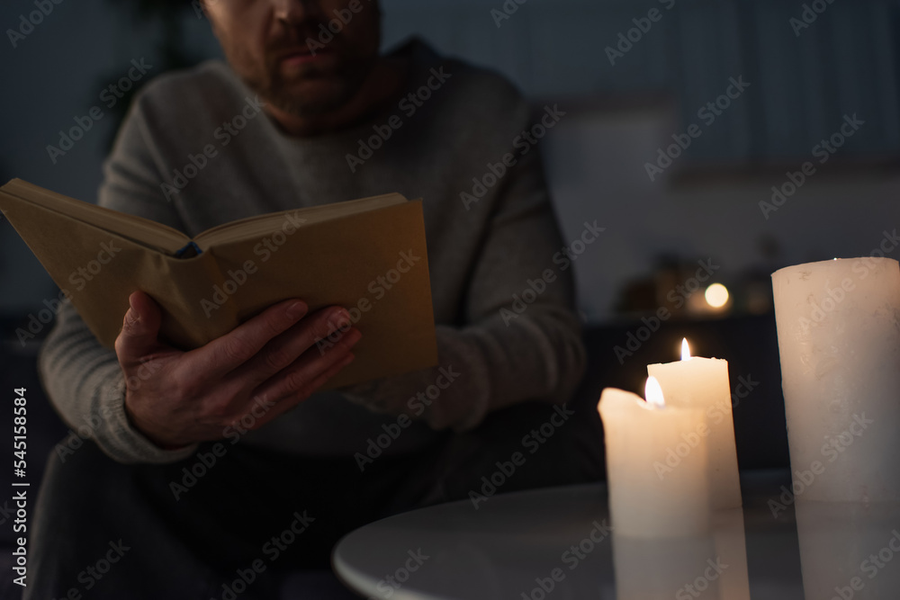 cropped view of man reading book near candles burning in dark kitchen  during power outage. Stock Photo
