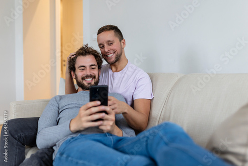 Cute young gay couple video calling their friends in their living room at home. Two male lovers smiling cheerfully while greeting their friends on a smartphone. Young gay couple sitting together.