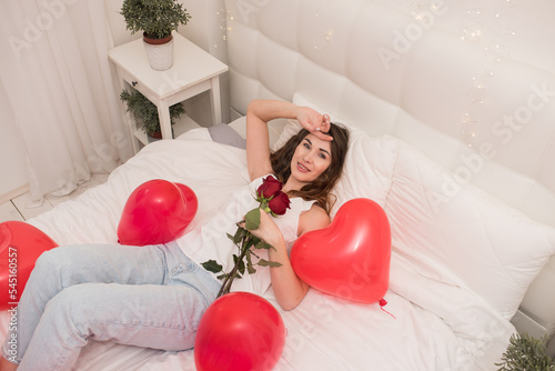 Valentine's day, birthday, red heart-shaped balloons, beautiful happy dark-haired girl lying on the bed with red roses in her hands