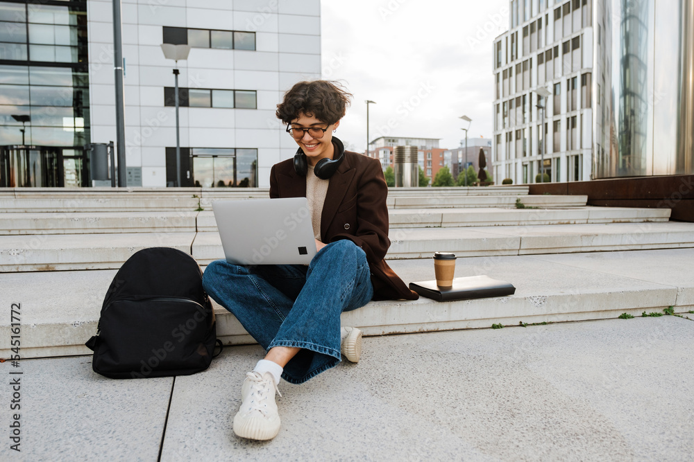 Young brunette woman using laptop while sitting on stairs over building Young brunette woman using laptop while sitting on stairs over building