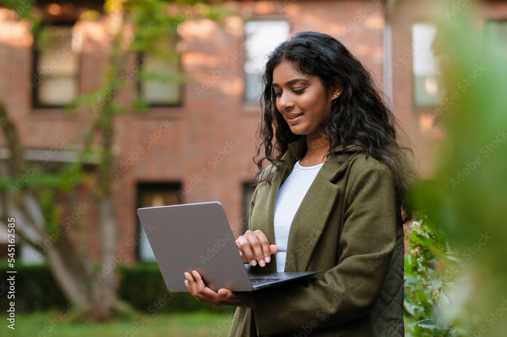 Brunette indian woman smiling while working with laptop on city street