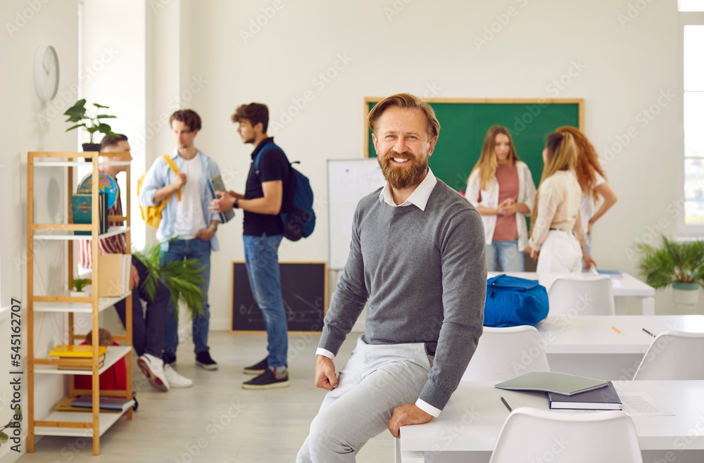 Portrait of a cheerful male teacher at work. Happy adult man smiling and looking at the camera while standing by a desk in the school classroom with students in the background Portrait of a cheerful male teacher at work. Happy adult man smiling and looking at the camera while standing by a desk in the school classroom with students in the background
