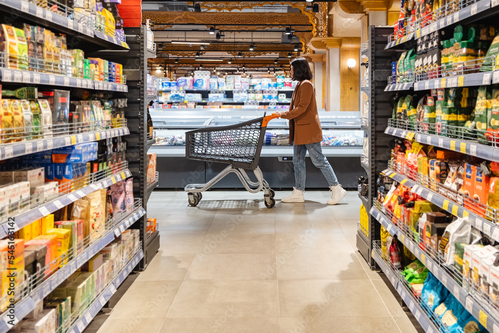 © phpetrunina14 - a woman with a cart walks between rows of shelves in a grocery store