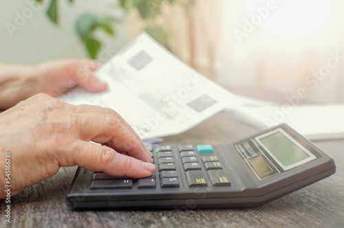 An elderly woman sitting at a table looks at bills for gas, water, electricity and counts on a calculator. Selective focus.