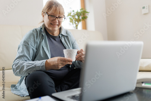 Serene mature female with blond hair having hot tea or coffee while working remotely in front of laptop at home