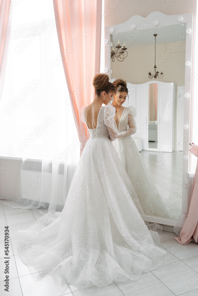a bride with a train of a wedding dress stands at the mirror with her back to the camera in a bright interior a bride with a train of a wedding dress stands at the mirror with her back to the camera in a bright interior