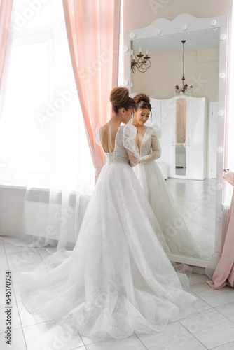 a bride with a train of a wedding dress stands at the mirror with her back to the camera in a bright interior
