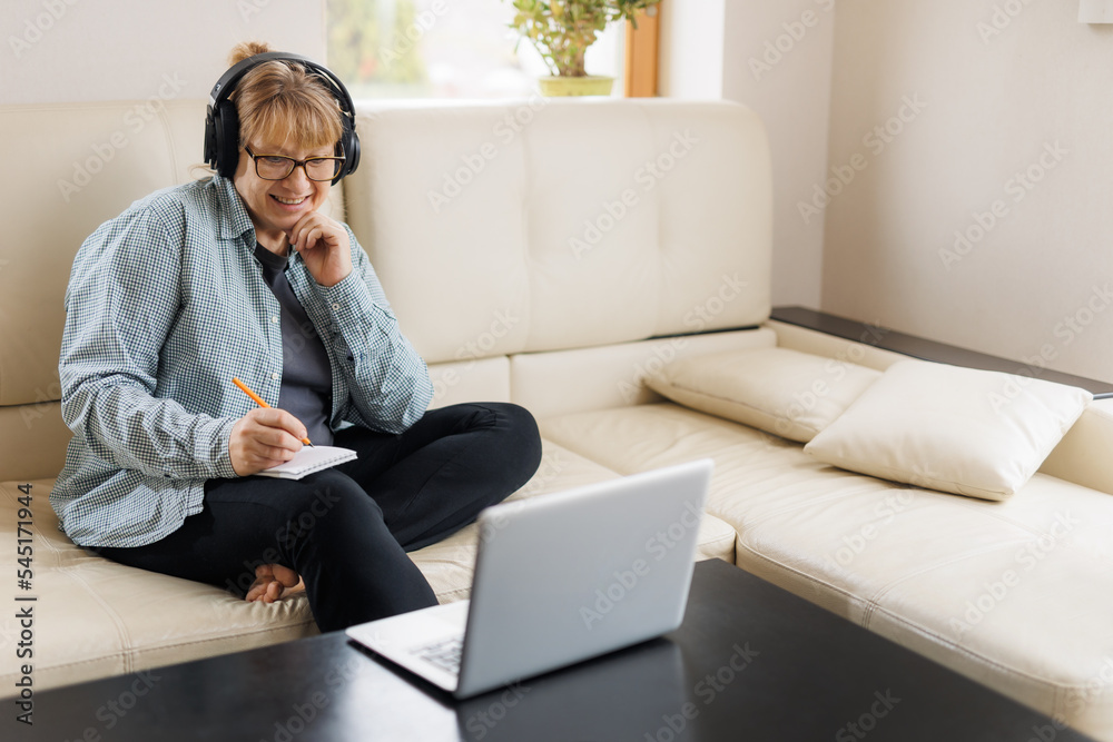 Modern middle aged blond woman in headphones take online course or training on computer at home. Smart mature Caucasian female in earphones watch webinar make note study distant on laptop.