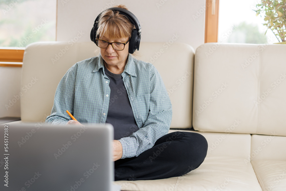 Modern middle aged blond woman in headphones take online course or training on computer at home. Smart mature Caucasian female in earphones watch webinar make note study distant on laptop. Modern middle aged blond woman in headphones take online course or training on computer at home. Smart mature Caucasian female in earphones watch webinar make note study distant on laptop.