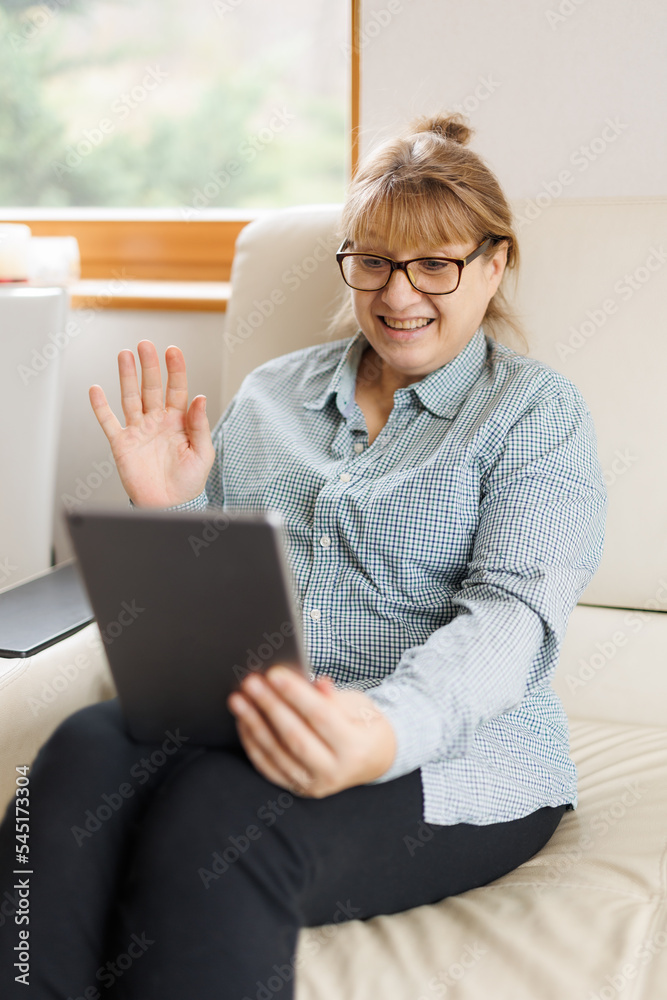 Fototapeta premium Beautiful mature woman in eyeglasses is using a digital tablet and smiling while sitting on couch at home