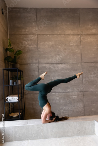 Young sporty attractive woman practicing yoga, doing headstand exercise, variation of salamba sirsasana pose, working out, wearing sportswear, grey top, indoor close up view, yoga studio, rear view